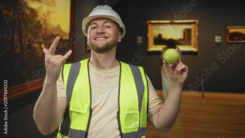 фотографія Man engineer in hardhat and high visibility vest holding green apple and making rock horns in building; playful mischief
