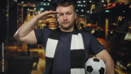 Foto Man salutes hand to forehead holding a soccer ball in a shop building with black and white scarf; pride fandom loyalty