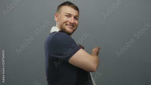 Fotografie Man with soccer scarf pointing with bare forearm and smiling while turning in gray studio set; cheerful mischief