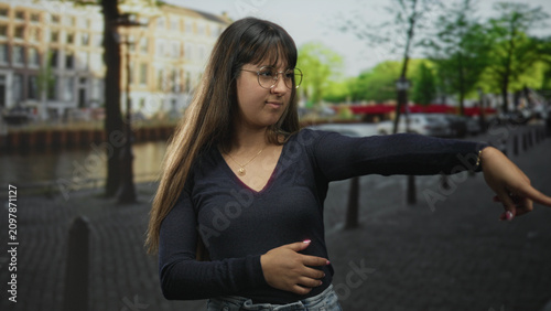 Konstfotografi Woman with glasses and long hair touches chin then extends arm and points finger along canal street with parked cars and bollards; curiosity