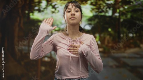 Canvas Print Young hispanic woman with hand raised and hand to chest making a pledge in a pink top on a tree-lined street; sincerity trust resolve