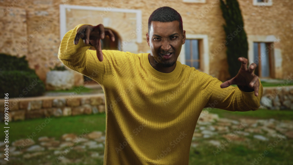 Naklejka premium Young man makes claw gesture with both hands while wearing a yellow sweater in front of a stone building, playful grin visible; playfulness energy.