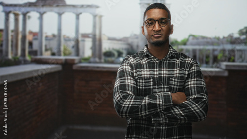 Canvastavla Young black man with glasses and arms crossed on building rooftop by brick railing, facing camera; confidence contemplation focus