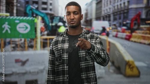Photos Young black man stands by a construction barrier on a city street, face visible and glancing sideways toward traffic; contemplation
