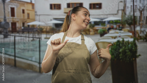 Woman in apron points thumb to herself and makes lively hand gestures in a busy street cafe setting; friendly confidence.