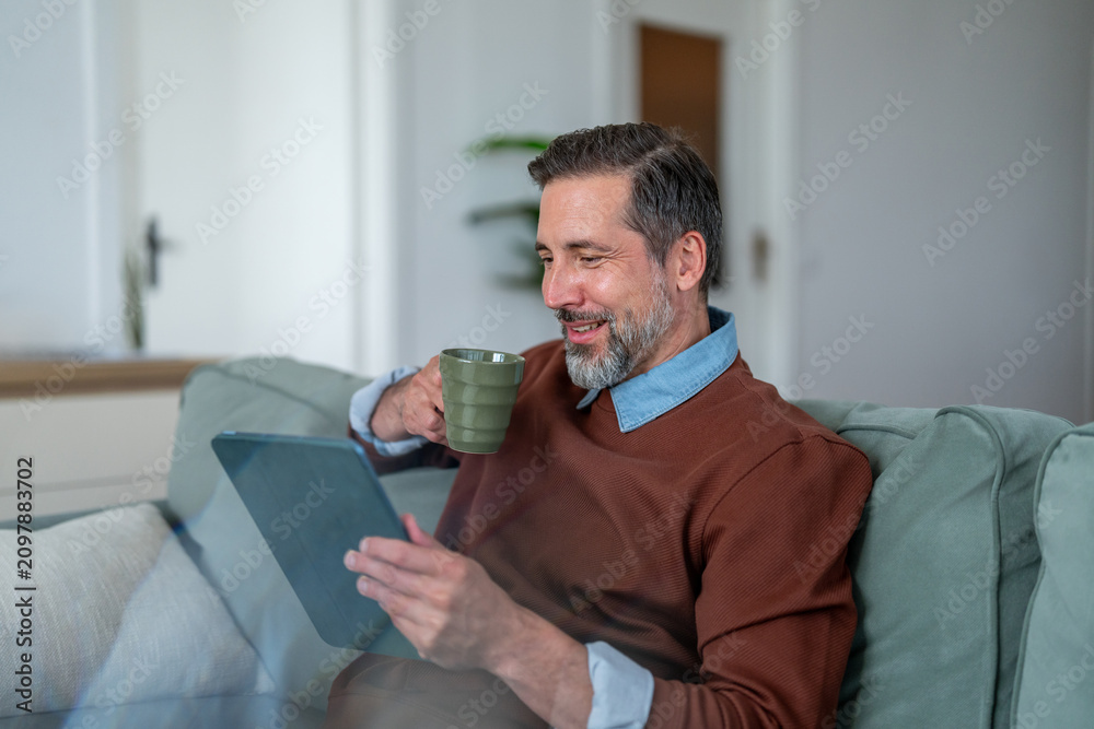 Fototapeta premium Smiling mature man relaxing on sofa using tablet drinking coffee