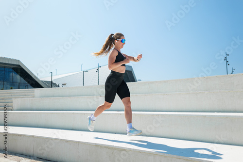 Running with energy under the bright sun, a woman showcases her fitness routine on a modern staircase