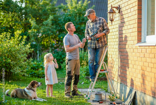 Senior man on a ladder installs a wall lamp while a younger man and little girl watch.
