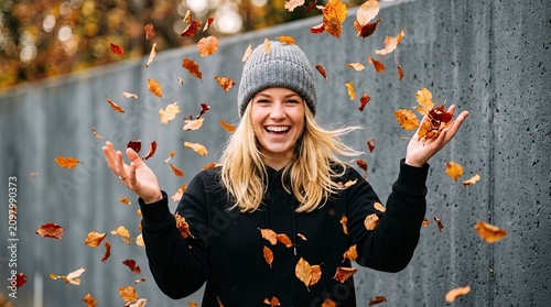 Young woman laughing joyfully while throwing autumn leaves in the air wearing a grey beanie and black hoodie against a textured concrete wall with conveying sense carefree happiness captures joyful