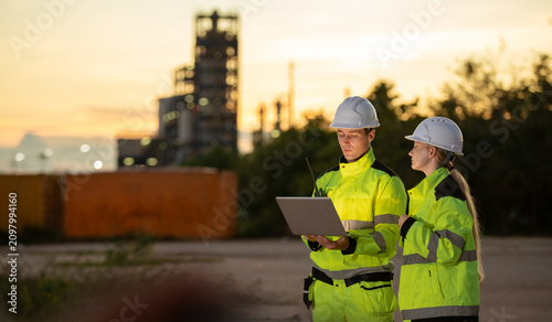 Engineers collaborating on construction site at sunset with safety gear and laptop in industrial environment