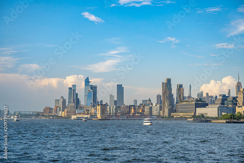 Midtown Manhattan skyline with Hudson Yards beside Hudson River. Ferries cross the water below the Empire State Building and waterfront towers under scattered clouds.