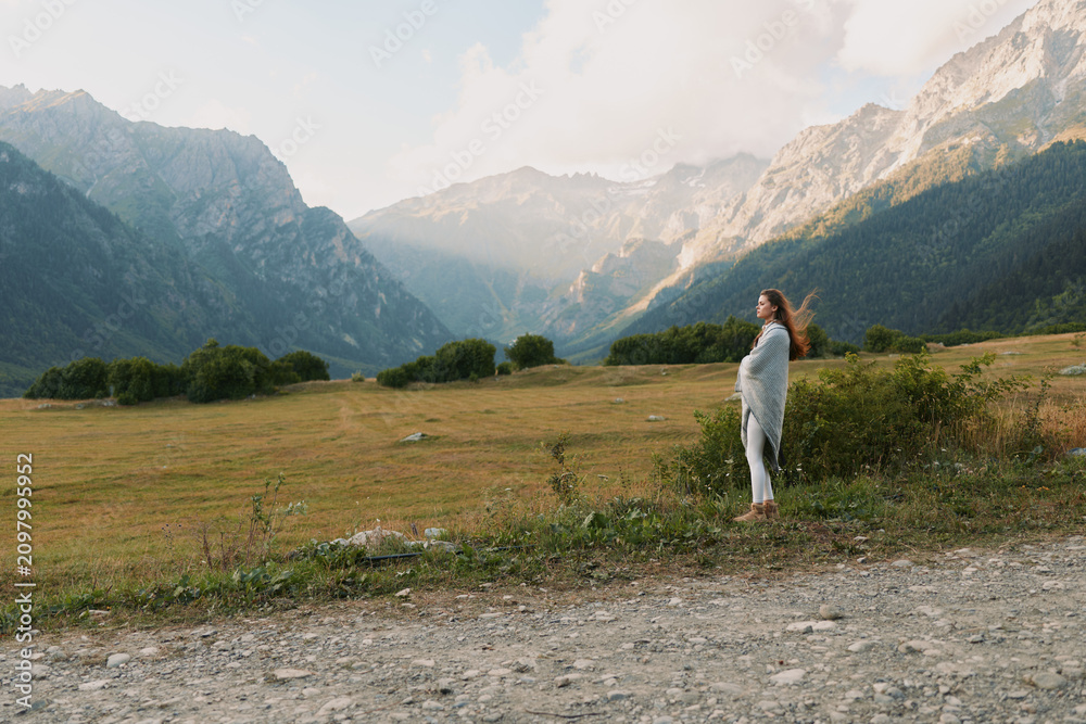 Obraz premium A woman in casual clothes stands on a dirt path in a vast mountain valley surrounded by green trees and rocky peaks under a cloudy sky. Lifestyle concept outdoors.