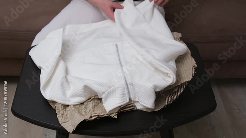 Person seated on brown sofa folding and packing striped clothing on small black table in calm home interior with soft natural light, neutral cushions, beige floor, and centered domestic composition