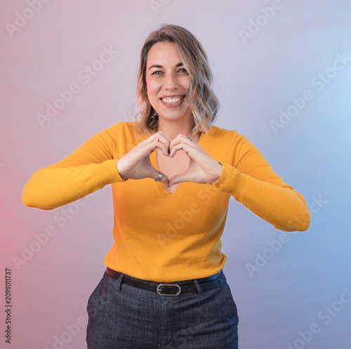 Smiling woman making a heart shape with her hands against a colorful studio background, representing love, care, kindness, gratitude and emotional connection.