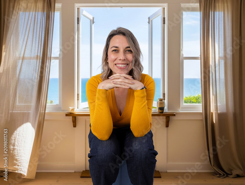 Smiling woman sitting by an open window at home, representing wellbeing, comfort, relaxation, positive lifestyle and enjoying a peaceful moment indoors.