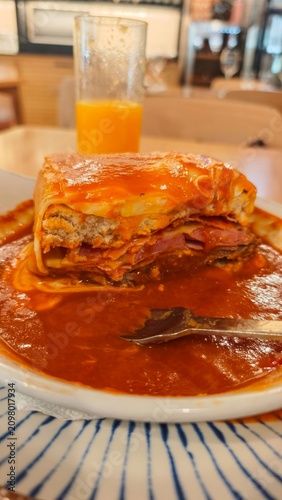 Layered lasagna with meat, cheese, and sauce served on a plate, accompanied by a glass of orange juice in a casual dining setting