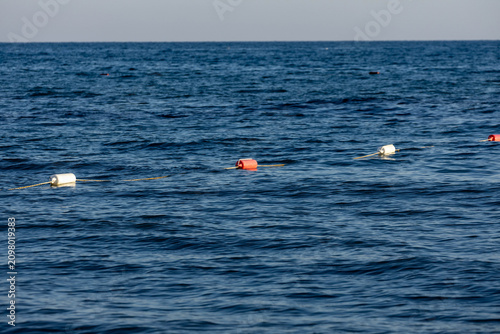 Bright red and white safety buoy on yellow rope barrier floating on dark blue sea water. Beautiful mediterranean beach boundary marker on calm sunny day in may, minimal landscape.