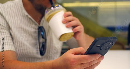 Man drinking iced coffee while browsing smartphone at cafe table, holding takeaway cup and checking phone in casual closeup with sunglasses hanging on striped shirt, mobile detail, hand texture