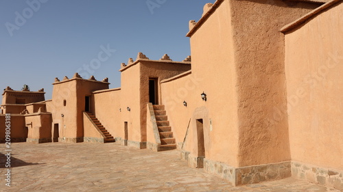 Sunlit adobe buildings with exterior staircases line a quiet stone courtyard