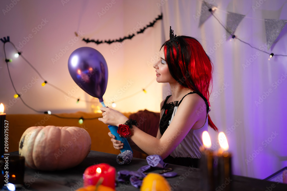 Fototapeta premium Cute Little Girl In A Halloween Witch Wig Blows Up A Balloon While Decorating A Holiday Room