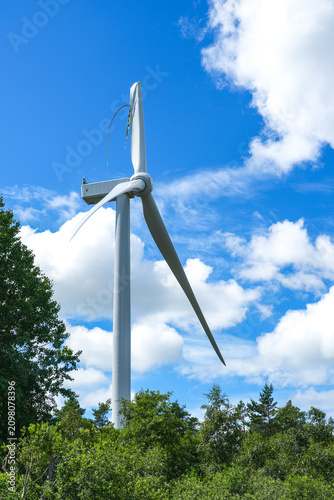 Damaged Wind Turbine with Broken Blade Rising Above Forest Under Bright Blue Sky and White Clouds View