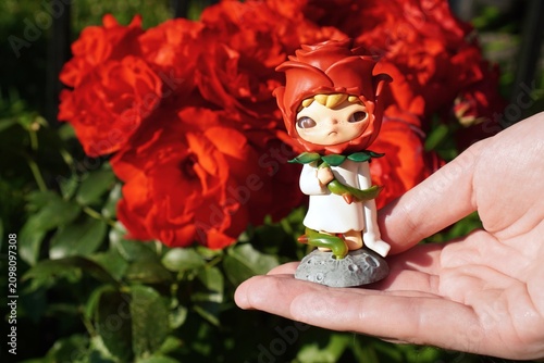 Charming figurine depicting a child in a rose petal outfit cradled gently in a hand. Vibrant red flowers in the background enhance the whimsical garden theme