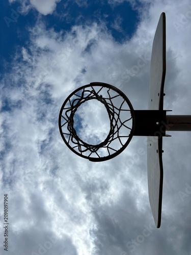 Low angle view of a basketball hoop and backboard silhouetted against a dramatic blue sky with fluffy white clouds. Concept of ambition, achieving goals, outdoor sports, and street basketball.