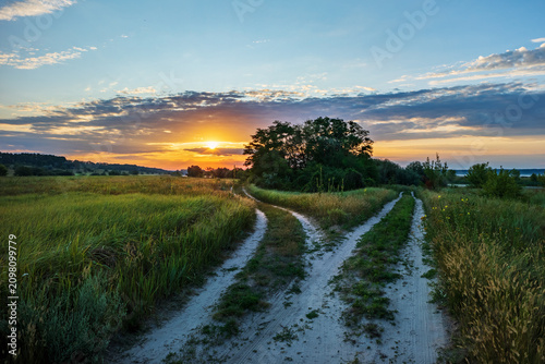 Sunrise over a meadow and country roads. Golden sunlight falls on the tall grass on a summer morning. A picturesque landscape of the forest-steppe natural zone