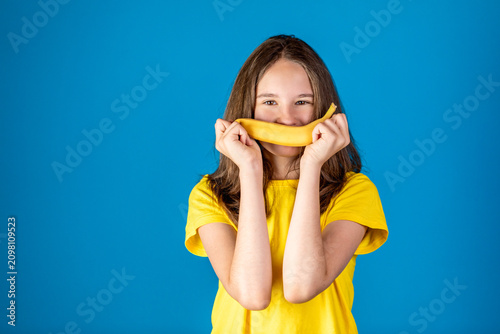 Playful young girl holding a banana in front of her mouth like a smile, wearing a yellow t-shirt against a blue studio background