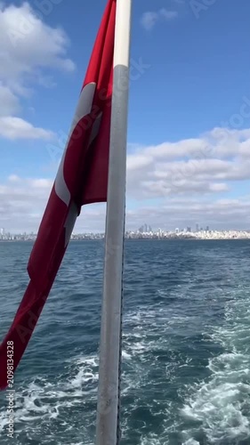 Turkish National Flag waving on the ferry boat headed through Bosphorus to Prince Islands. Cloudy sky and wavy sea. The distant view of Istanbul cityscape 