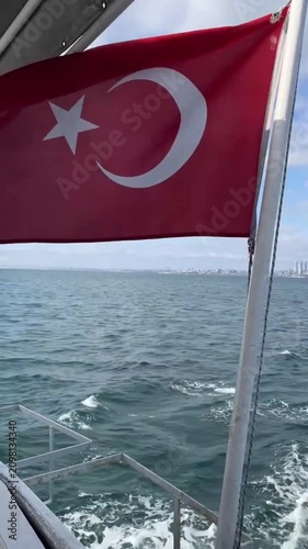 Turkish National Flag waving on the ferry boat headed through Bosphorus to Prince Islands. Cloudy sky and wavy sea. The distant view of Istanbul cityscape 