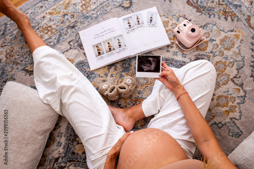 Top down view of a pregnant woman holding an ultrasound scan photo beside an open pregnancy album an instant camera and knitted booties on a rug a tender maternity scene.