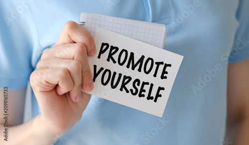 Hand holds paper with bold text: PROMOTE YOURSELF. Person wears light blue shirt, business concept