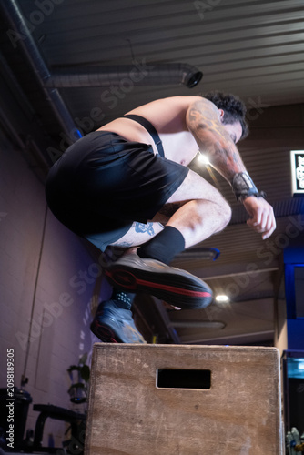 man jumping over wooden box in indoor gym with suspension straps, energetic focused workout scene, black brown white red colors, low angle shot from below the box.