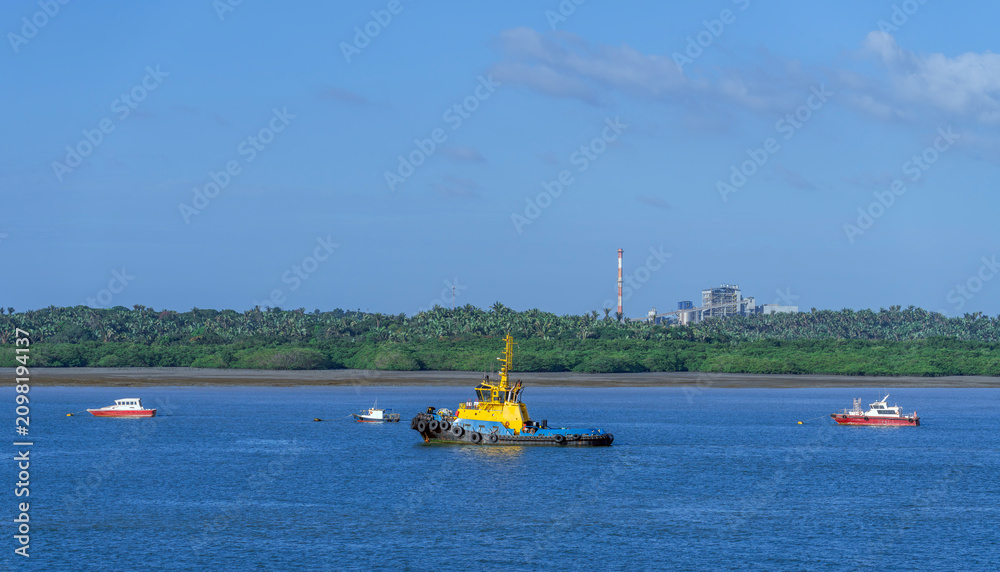 Fototapeta premium Vibrant yellow and blue tugboat navigates calm blue waters alongside smaller vessels. The background reveals a lush green shoreline contrasting with an industrial plant and tall smokestack