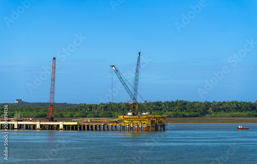 Industrial marine construction site featuring a heavy yellow pier structure with tall lattice boom cranes over blue water. A dense green coastal forest forms the background