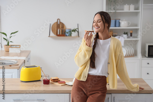 Konstfotografi Smiling young woman enjoying a piece of toast with jam in a bright modern kitche