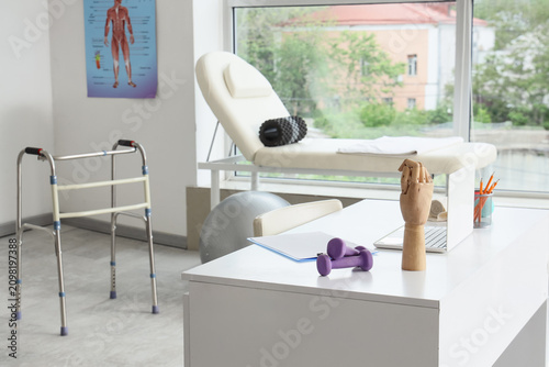 Physical therapy room with a walker, examination couch, and desk with dumbbel...