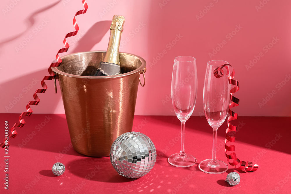 Fototapeta premium Disco balls and ice bucket with bottle of champagne on red table near pink wall