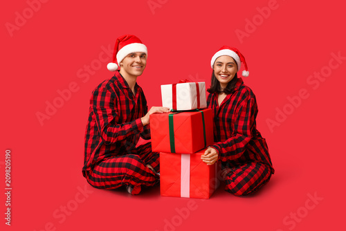 Happy young couple in Christmas pajamas and with gifts on red background