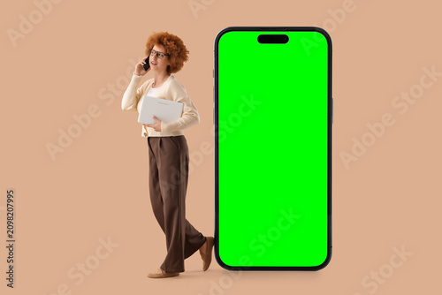 Young woman with laptop talking on phone next to large smartphone with green screen  digital communication and technology