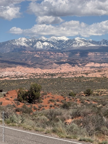 Snow capped La Sal Mountains rising above red sandstone dunes and desert scrub in Arches National Park Utah
