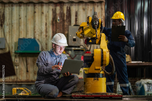 Worker in a white hard hat sits with a laptop. Another worker in a yellow hard hat holds a tablet. Yellow robotic arm stands in an industrial workshop. Metal walls and equipment fill the background