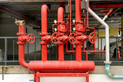 Red industrial fire protection pipes featuring valves and pressure gauges in a parking garage. Professional fire sprinkler pipeline installation for building safety and emergency water response.