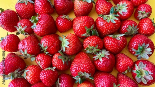 Horizontal banner of ripe red strawberries with green leaves arranged on a bright yellow background. Top-down view of organic summer berries pattern, wide frame template for website header.