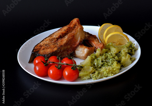 Photos Two grilled chum salmon steaks, cherry tomatoes, and boiled broccoli on a round plate against a black background