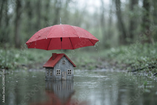 Miniature wooden house protected by a red umbrella in a rain puddle