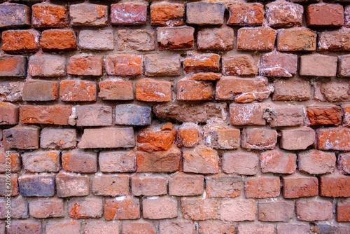 Detailed view of a rustic brick facade featuring a mix of orange, red, and pale bricks, with visible damage, crumbling joints, and a peeling paint residue
