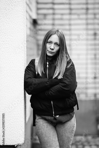 Black and white portrait of a young serious woman in jacket leaning against a textured stone wall in an urban underpass, looking at camera.