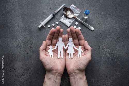 Protective hands holding a paper cut-out family surrounded by drug paraphernalia, symbolizing the impact of addiction on family life and safety.